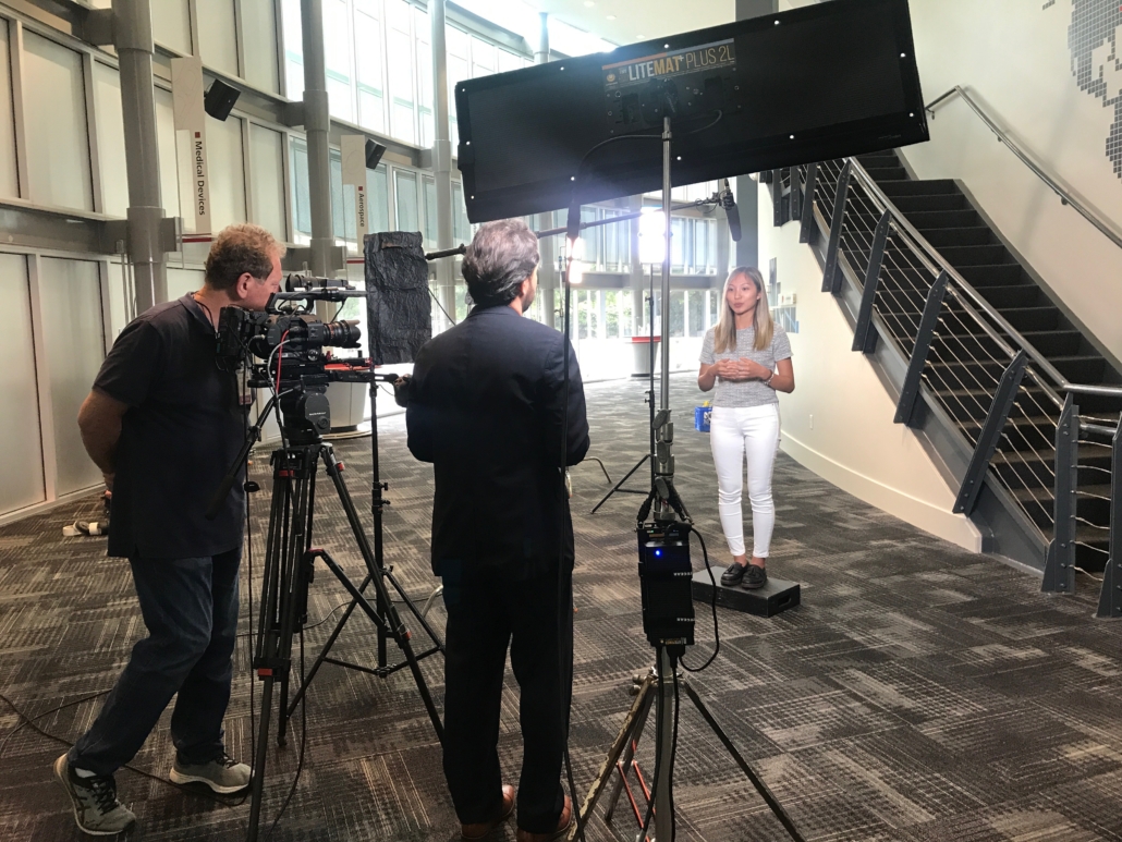 Two crew members behind a tripod-mounted camera filming a presenter speaking to camera in a building atrium